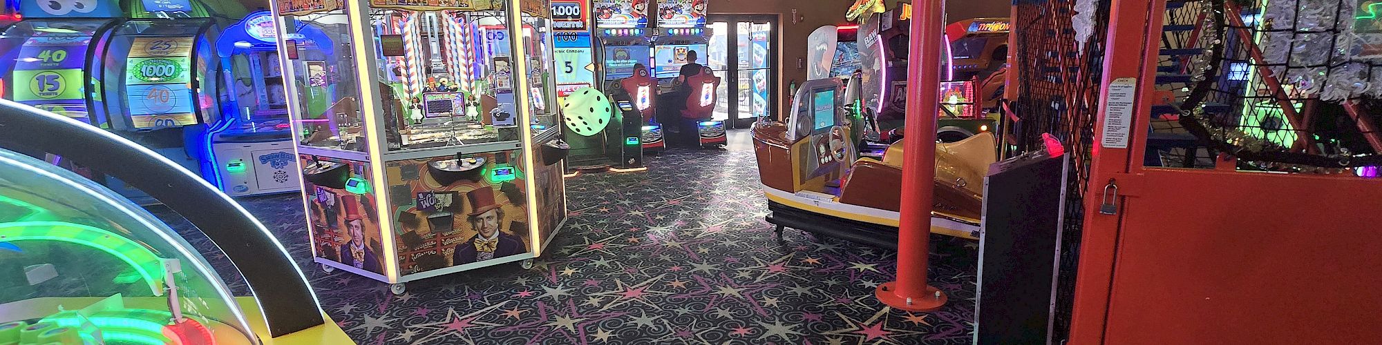 An arcade full of colorful claw machines, prize games, and neon lights under a starry carpet pattern, with balloons and bright, upbeat decor.
