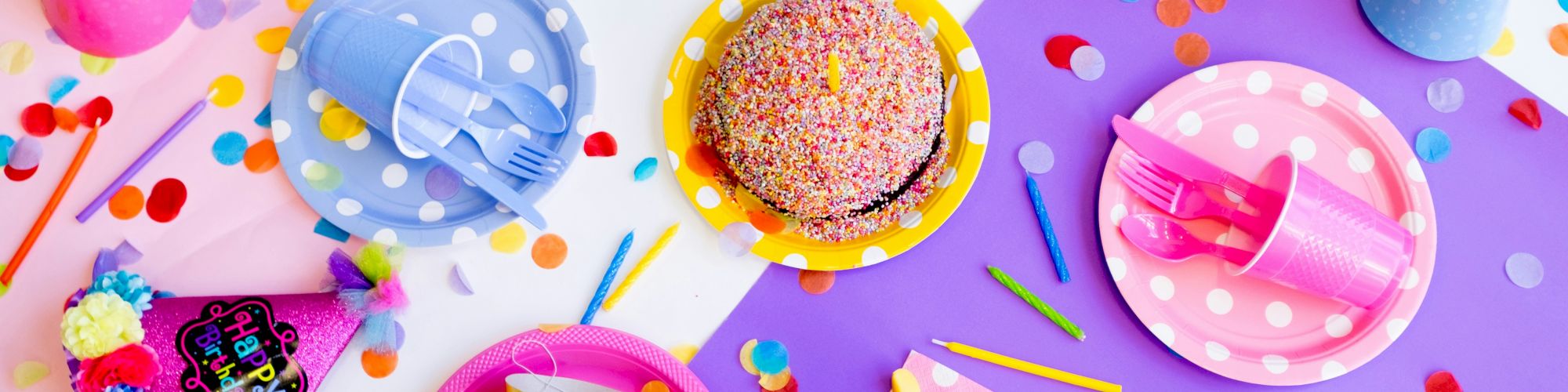 Colorful birthday setup with party hats, polka dot plates, cups, a sprinkles cake, and confetti scattered on a white background.