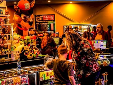 Kids and a woman at a colorful arcade or toy shop, with a large hanging plush and shelves of figures in the background.