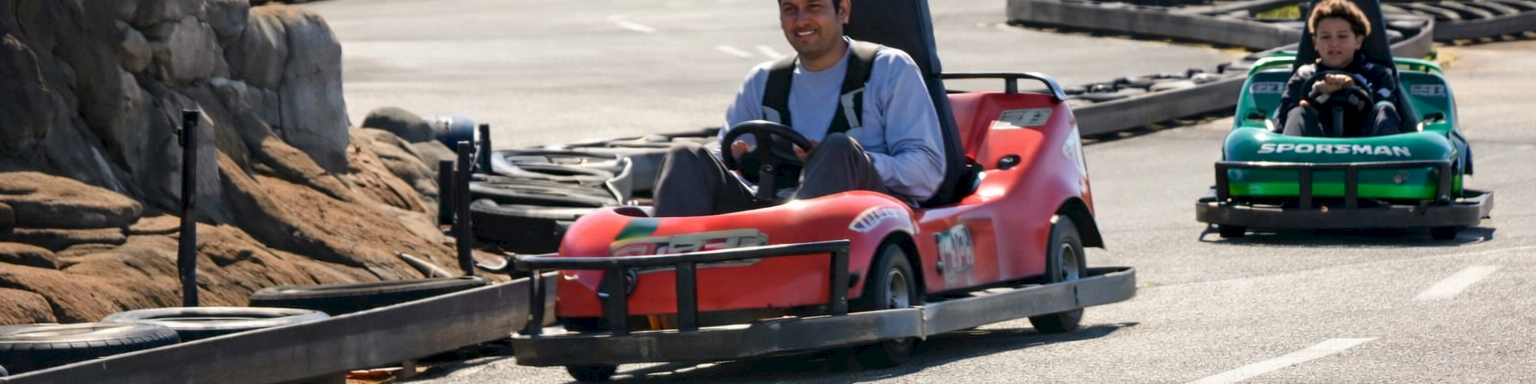 Two people race go-karts on a paved track, one red with a helmeted driver in front and a green kart behind in a parking-l lot area.