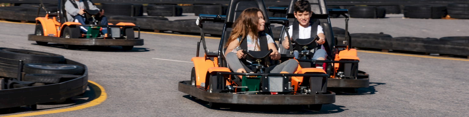 Three people in go-karts race around a track on a sunny day, navigating a curved section with a fence and trees in the background.