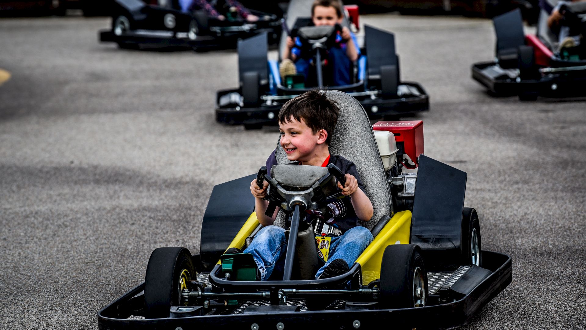 Children driving go-karts on a track, with a young boy in the closest kart smiling and looking ahead, enjoying the activity.