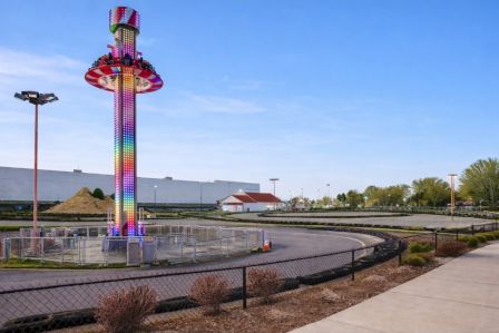 A colorful amusement ride stands by a fenced circular track; a bright ride tower glows, with a clear blue sky and a wide empty parking lot nearby.