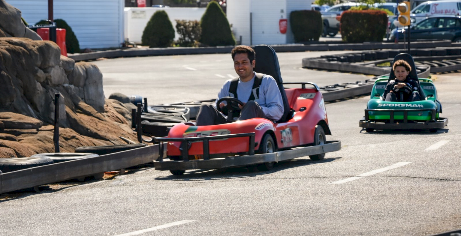 Two people race pink and green go-karts on a track, with a rocky barrier and course signage in the background.