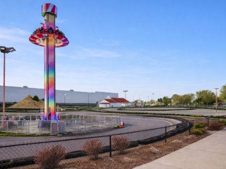 An outdoor amusement ride tower with colorful LED lights rises above a fenced fairground as evening light fills the sky.