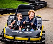 A woman and a child ride a small “Tiny Scorpion” go-kart together on a paved track.