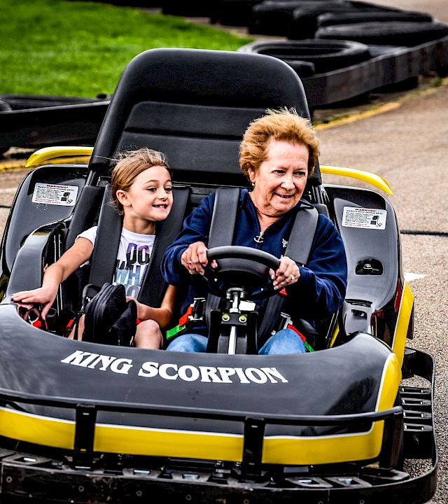 A woman and a child ride a small “Tiny Scorpion” go-kart together on a paved track.
