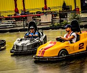 Two drivers smile in race go-karts on an indoor track under bright yellow curtains.
