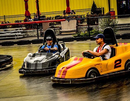 Two drivers smile in race go-karts on an indoor track under bright yellow curtains.