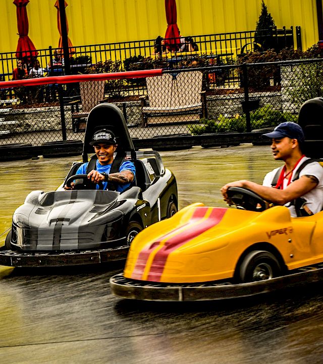 Two drivers smile in race go-karts on an indoor track under bright yellow curtains.