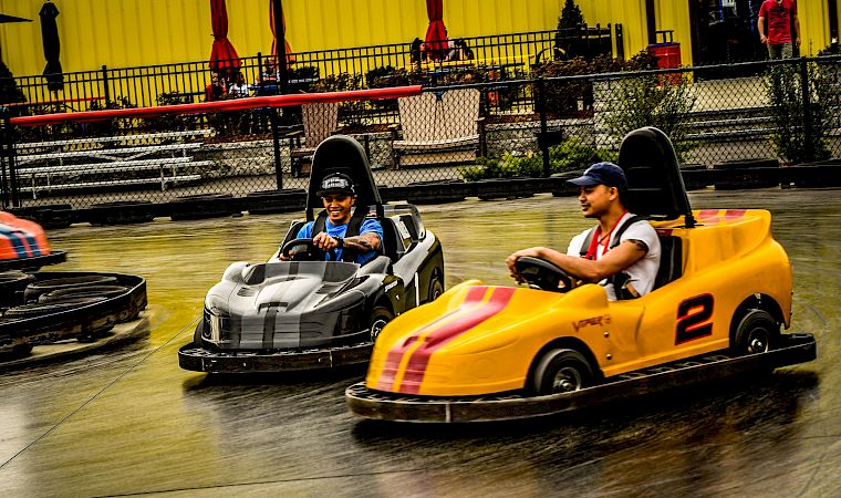 Two drivers smile in race go-karts on an indoor track under bright yellow curtains.