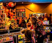 At a carnival arcade ticket booth, a fox-like mascot and plush toys sit by prize counters as people queue at neon signs.