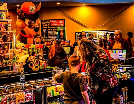 At a carnival arcade ticket booth, a fox-like mascot and plush toys sit by prize counters as people queue at neon signs.