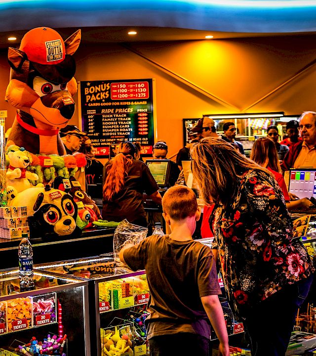 At a carnival arcade ticket booth, a fox-like mascot and plush toys sit by prize counters as people queue at neon signs.