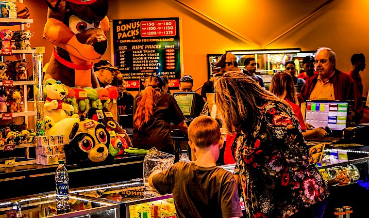 At a carnival arcade ticket booth, a fox-like mascot and plush toys sit by prize counters as people queue at neon signs.