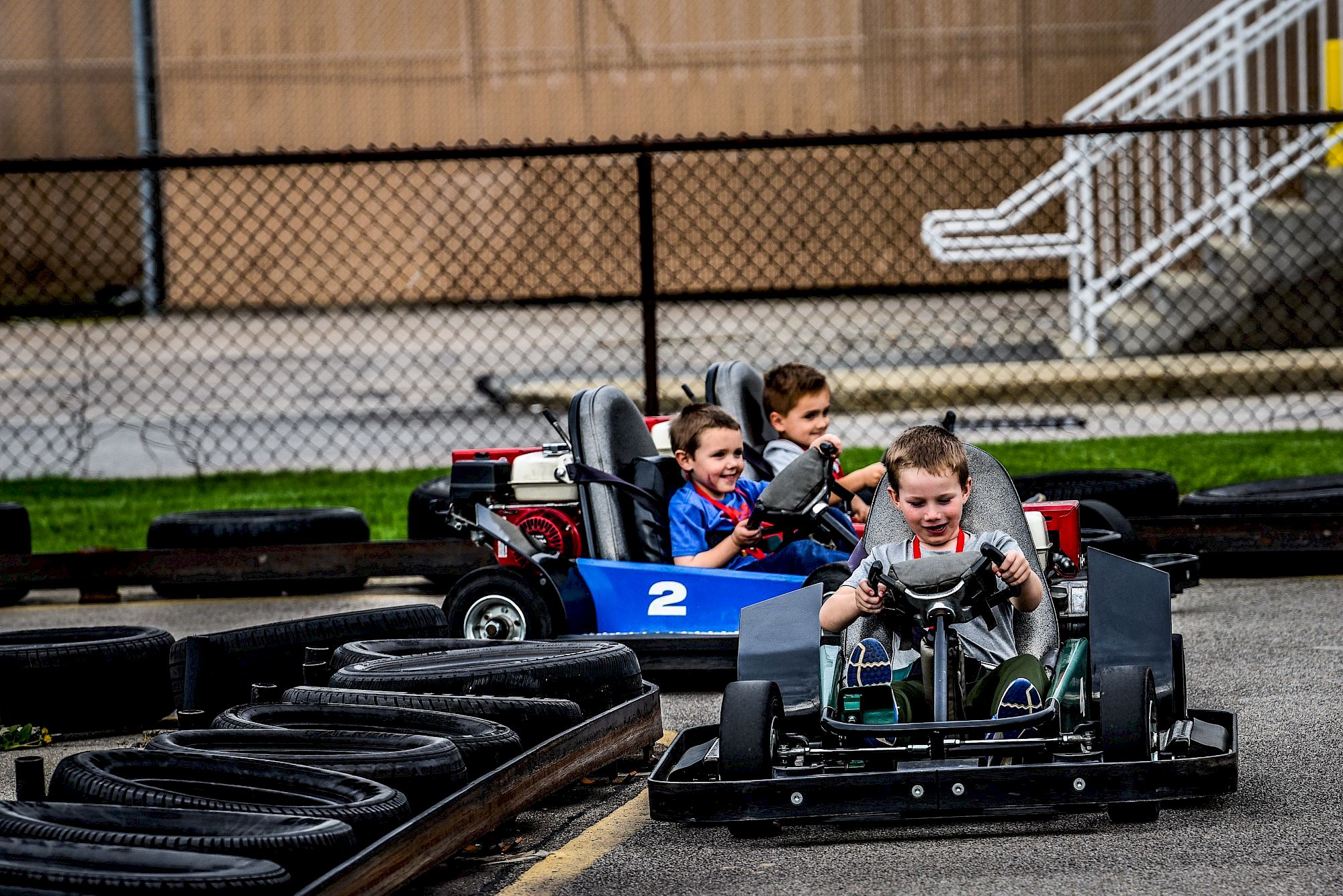 Kids ride go-karts on a race track behind a chain-link fence, gripping steering wheels and smiling.