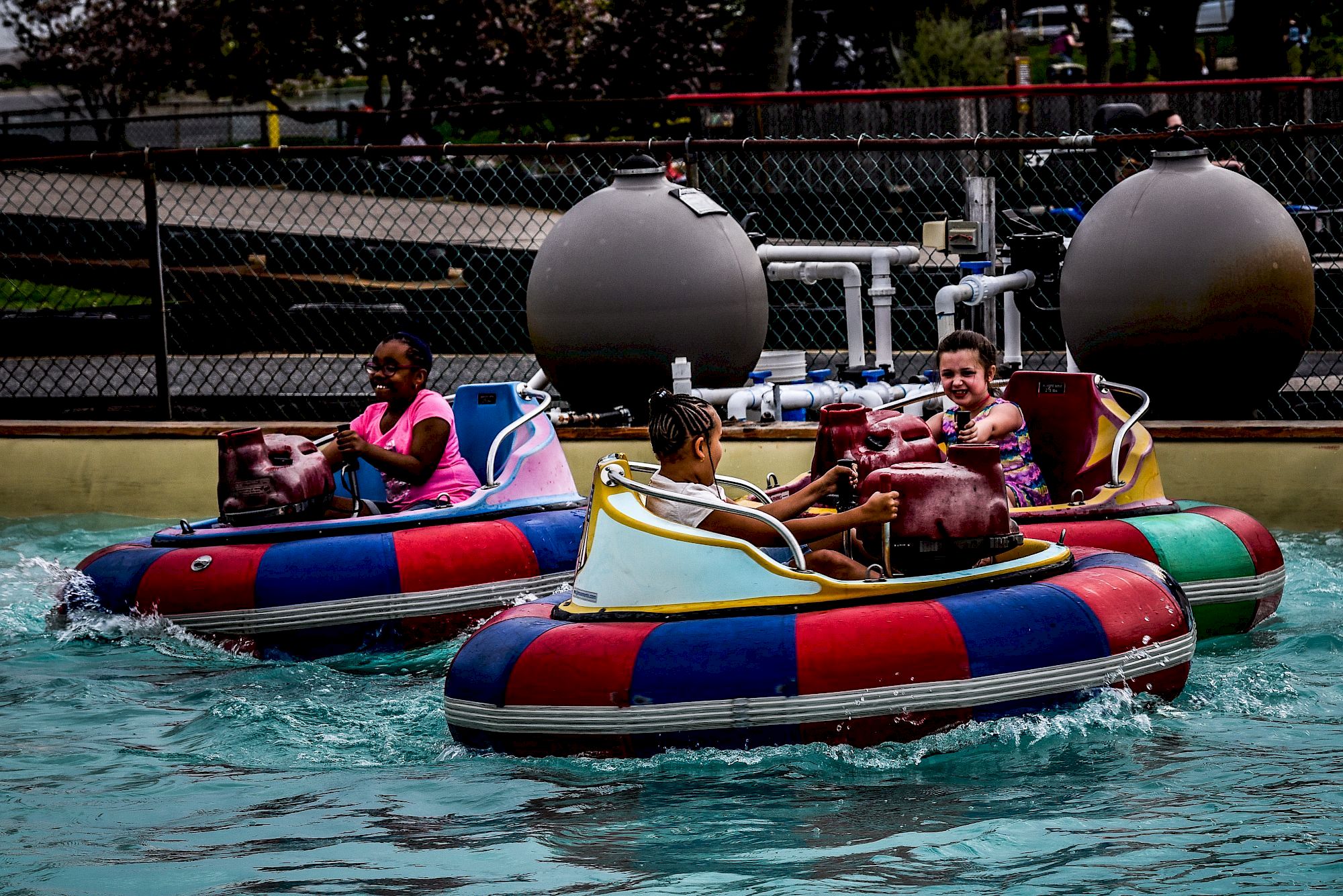Two kids ride colorful inflatable bumper boats on a pool in an amusement park, splashing through the water.