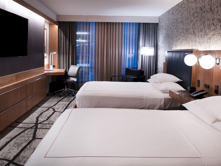 The image shows a modern hotel room with two queen beds, a wall-mounted TV, a desk, a chair, and a large window with curtains.