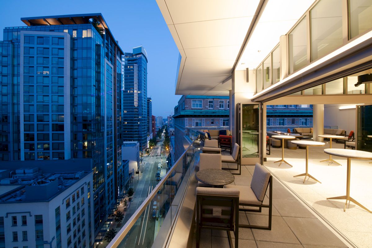 A modern urban balcony at dusk with outdoor seating offers views of high-rise buildings and a busy city street below.