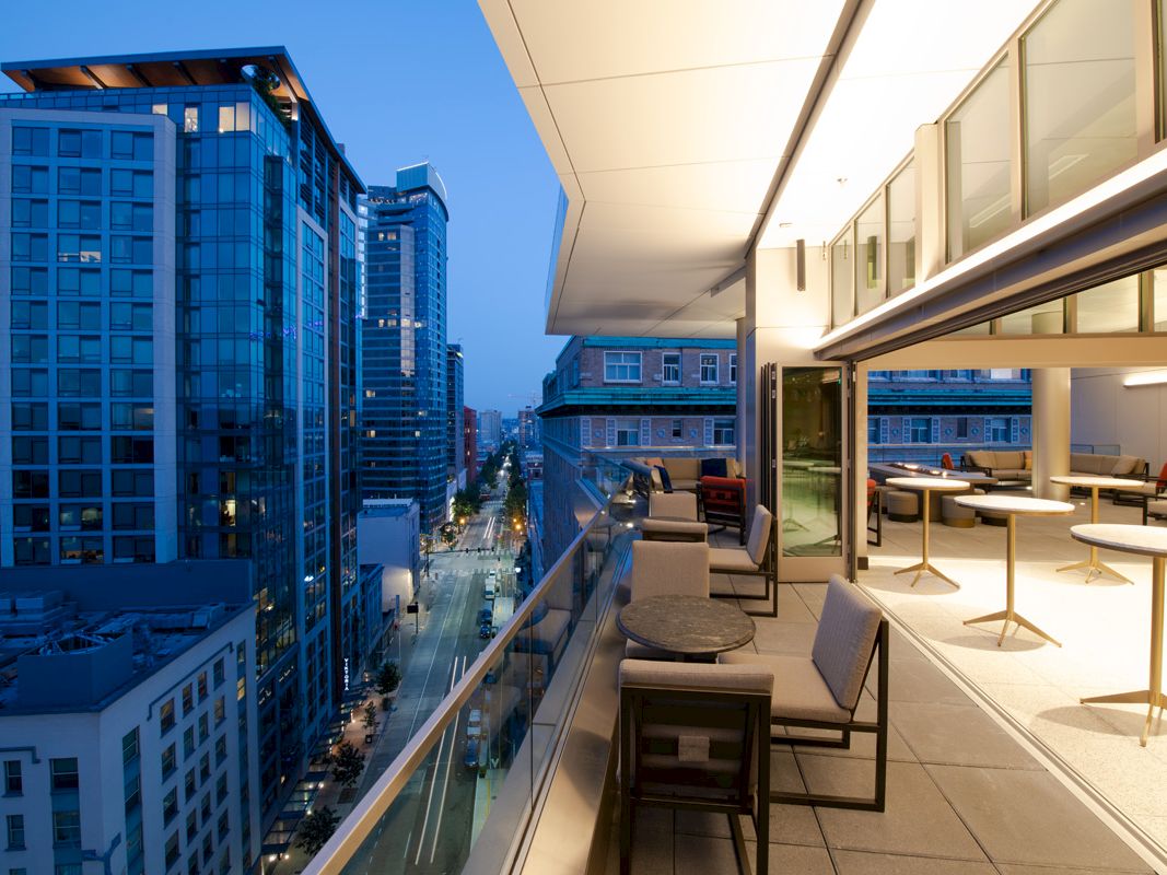 A modern urban balcony at dusk with outdoor seating offers views of high-rise buildings and a busy city street below.
