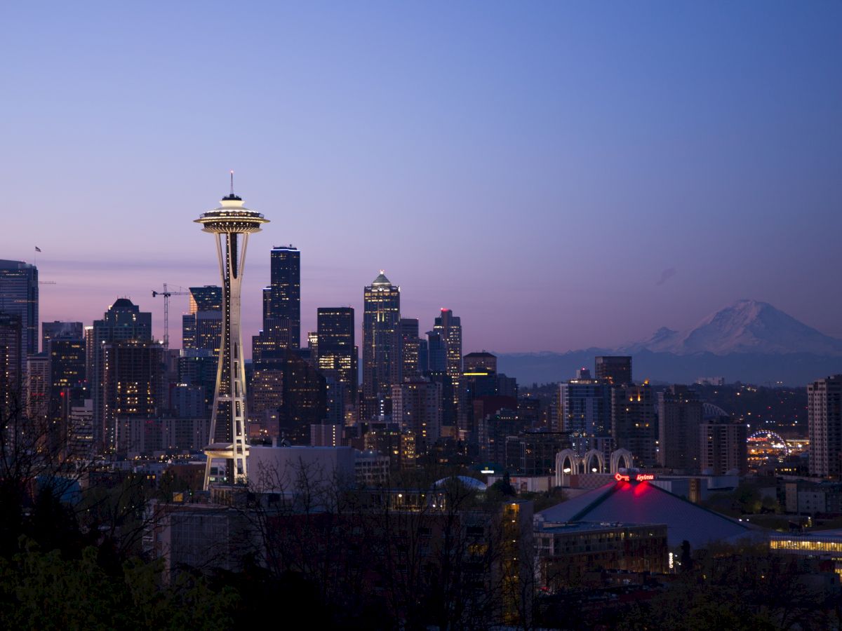 A city skyline at dusk featuring a prominent tower structure, with Mount Rainier visible in the background under a clear sky.