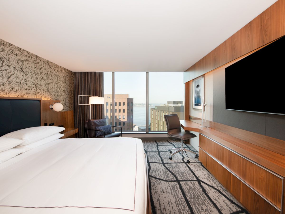 A modern hotel room featuring a large bed, a flat-screen TV, a work desk with a chair, and a window with a city view in the background.