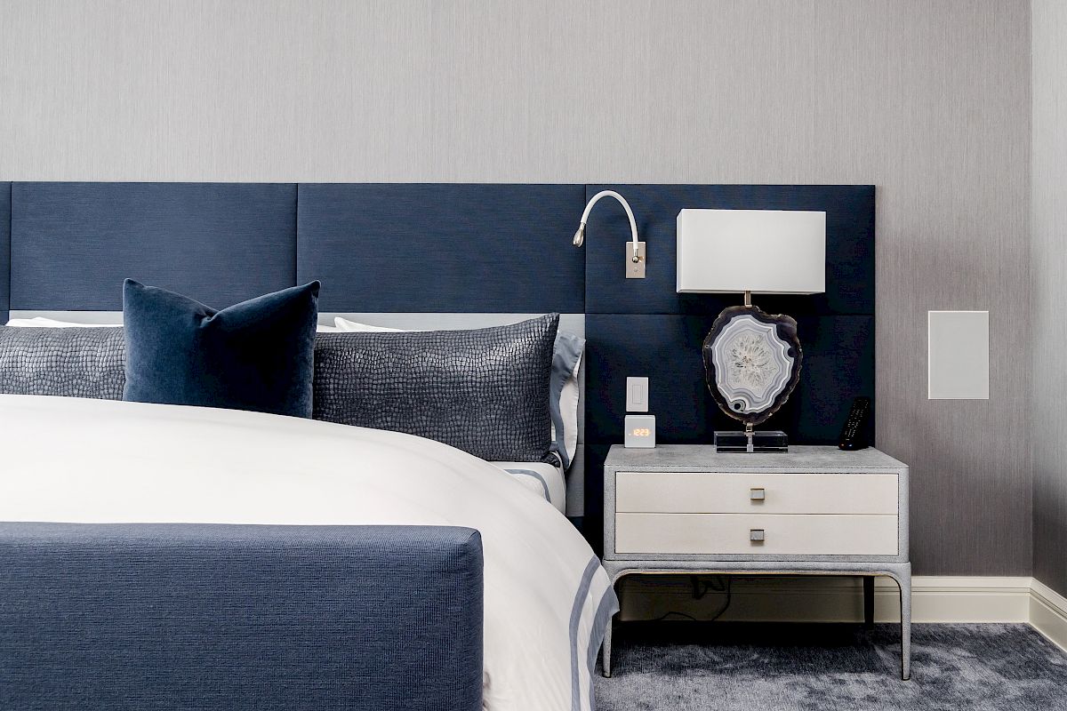 A modern bedroom with a blue headboard, white bedding, bedside table, lamp, and decorative items. The room has a clean and minimalist design.
