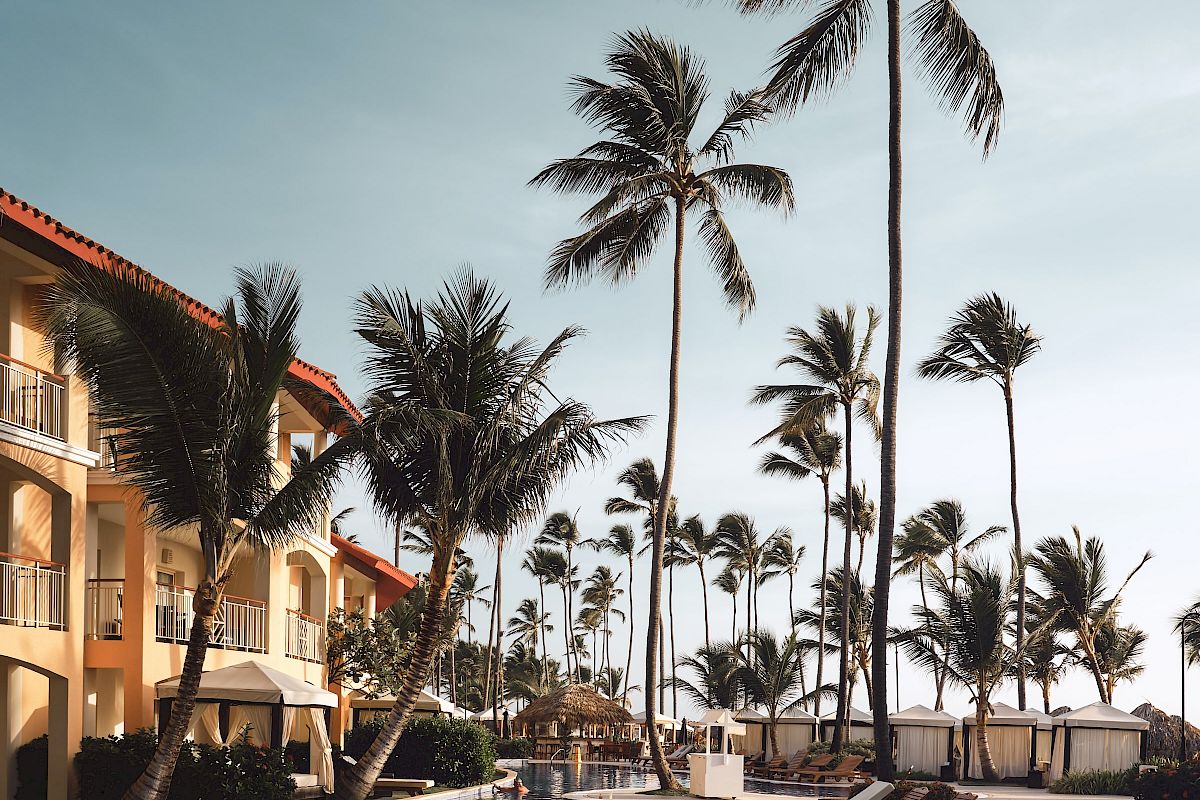 A serene poolside scene with lounge chairs, a pool, tall palm trees, and a resort building under a clear sky during daytime.
