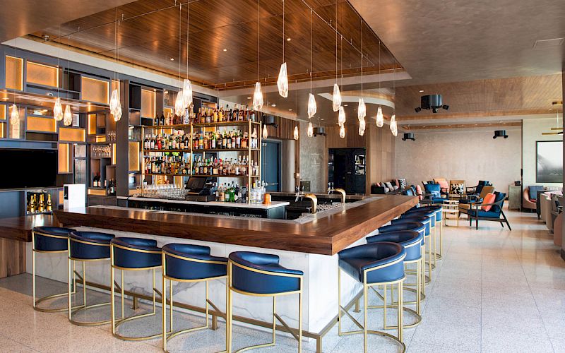 This image shows a modern bar with a wooden countertop, blue stools, hanging lights, and a well-stocked bar in the background, with seating areas nearby.