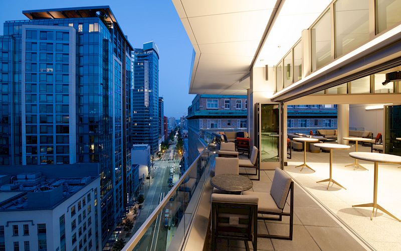 A modern balcony with seating areas overlooks a cityscape of tall buildings and streets illuminated in the evening.